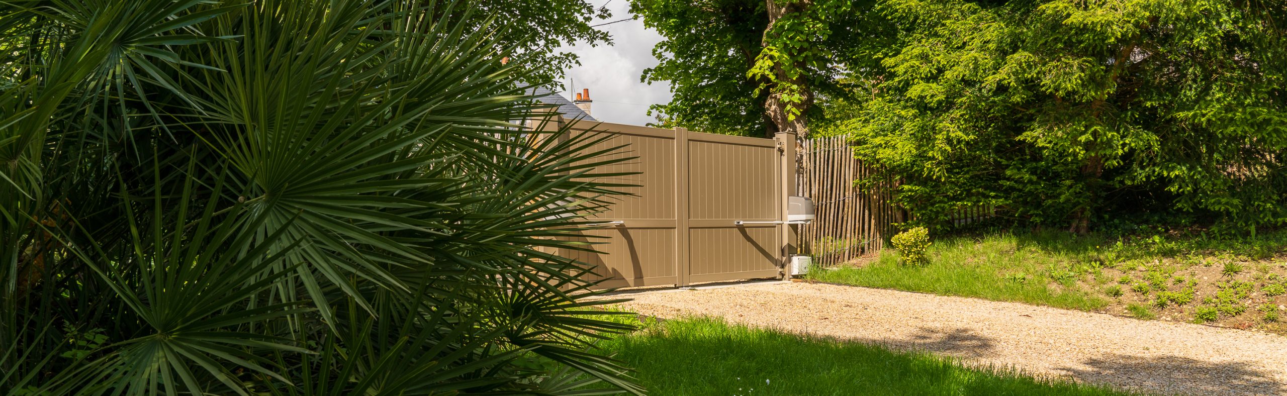 Allée de jardin avec portail battant motorisé beige, palmiers et végétation luxuriante